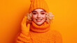 © Shutter2U - A cheerful young woman in a cozy oversized orange sweater, matching beanie, and gloves stands against a vibrant orange background, radiating warmth and joy. Perfect for autumn fashion themes.