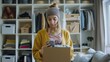 © Milan - A young woman in a cozy modern room, wearing a yellow cardigan and knit headband, holds a box while looking at her smartphone, implying organization and multitasking.
