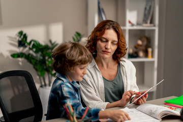 in the children's room a schoolboy in a white sweater and a blue checkered shirt at desk does homework mom explains the task