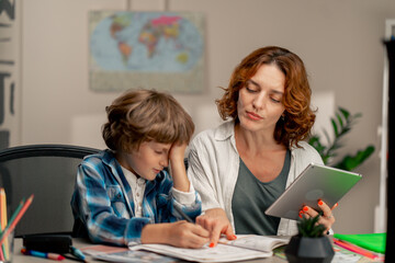 in the children's room a schoolboy in a white sweater and a blue checkered shirt at desk does homework mom explains the task