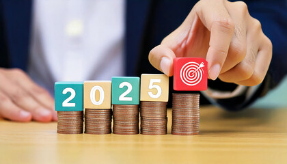 A hand places a target block on stacked coins marked with the year 2025, symbolizing financial goals and future planning.