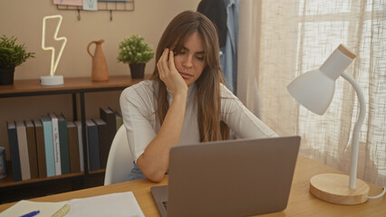 A thoughtful young woman working on a laptop in a well-lit home office setup