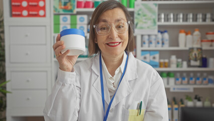 Mature woman pharmacist holding cream in a drugstore showcasing healthcare products.