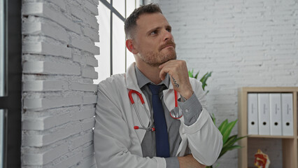 A thoughtful man with a beard wearing a lab coat and stethoscope poses in a clinic office.