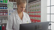 © Krakenimages.com - A young caucasian woman pharmacist working in a drugstore, focused on a computer monitor with shelves of products in the background.
