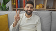 © Krakenimages.com - A smiling hispanic man making a peace sign while sitting on a grey sofa inside a cozy living room.