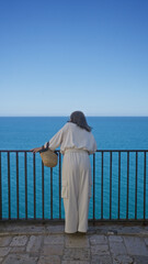 Wall Mural - A young hispanic woman gazes at the expansive ocean, standing on a viewpoint in polignano a mare, puglia, italy, under a clear blue sky.