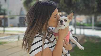 Wall Mural - A beautiful young hispanic woman kisses her chihuahua dog in an urban park setting, showcasing a loving bond between a pet and its owner in an outdoor city environment.