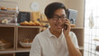 © Krakenimages.com - Young chinese man in a bakery smiling while talking on his phone surrounded by pastries and bread on display.