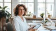 © Prostock-studio - A professional woman sits at a well-organized office desk, holding a tablet. Sunlight streams through large windows, illuminating plants and stationery, creating a bright and inviting workspace.