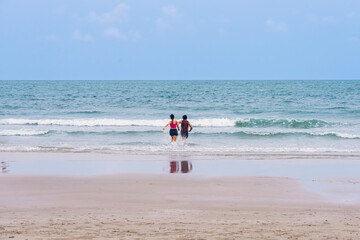 Back view of happy carefree couple holding hands and running in water at beach. Couple plays in ocean waves. Happy young couple playing in sea water. Guy and girl splashing together, beach vacation.