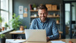 © Chahal - A middle-aged Caucasian man with short brown hair and a beard, wearing a blue shirt, smiling while working on a laptop computer on a desk in a home office setting