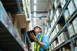 © KANGWANS - A man in a yellow vest is looking at a tablet while standing in a warehouse. He is wearing a hard hat and safety glasses