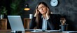 © Chayan - A woman in a business suit shows signs of stress while working at her desk, looking overwhelmed in a professional office setting.