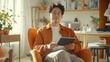 © Prostock-studio - A young man with curly hair and glasses sits comfortably in a bright, orange armchair, holding a tablet.