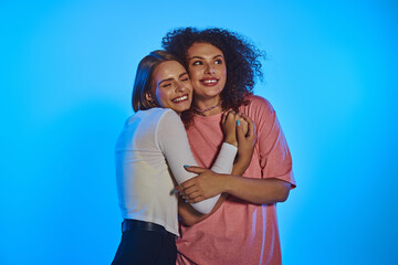 Wall Mural - Two joyful women embrace warmly, showcasing their love against a colorful backdrop of pink.