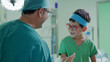 © Gita - smiling doctor in scrubs engages with a cheerful child wearing red glasses and a headband in a hospital setting, highlighting trust and positive patient care.