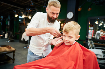 Barber using comb and shaver to cut hair. Professional hairdresser shaving little kid's nape.