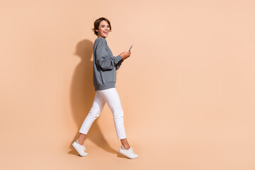 Full length photo of good mood girl wear pullover white trousers walk to empty space with smartphone isolated on beige color background