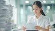 © Nano Photos - A woman in a white shirt focuses on paperwork at an office desk, surrounded by a tall stack of documents, highlighting a busy workday.