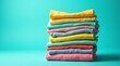 © Alex Pios - A neatly folded stack of colorful clothes stands against a vibrant turquoise backdrop, radiating joy and tidiness with each layer of bright fabric.
