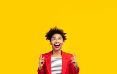 Look Here. Excited black girl pointing two index fingers up at copyspace isolated over yellow studio wall