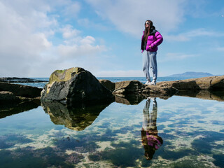 A girl in a purple jacket stands on stone shore. She is wearing gray sweatpants and. The ocean is in the background and the sky is cloudy and blue. Travel and tourism. Mullaghmore, Ireland.