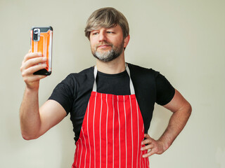 Butcher in black shirt and classic red and white stripe apron on light color background. Model in 40s with grey beard and hair. Meat industry worker with smart phone. Online advice concept.