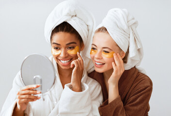 Home spa. Pleasant young multiracial young women putting eye patches and looking at mirror