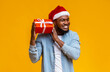 © Prostock-studio - Guess what's inside. Happy black man in Santa hat shaking Christmas present, listening what rattles in gift box, yellow background