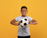 Cheerful black school boy holding soccer ball and smiling at camera, playing football, yellow studio background, copy space. Sport, leisure, entertainment, hobby for kids concept