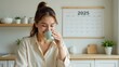 © 18042011 - A young woman smiling as she enjoys a cup of coffee in a cozy kitchen with a 2025 wall calendar behind her, symbolizing a fresh start to the new year with warmth, comfort, and personal goals