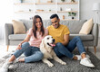 © Prostock-studio - Full length portrait of cheery multiracial couple with their pet dog sitting on floor at home. Arab man and his Caucasian wife spending time with golden retriever in living room