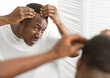© Prostock-studio - Dandruff Problem. Worried African American Man Looking At Flaky Scalp In Mirror, Touching Head Examining Gray Hair And Hairloss Issue Standing In Bathroom At Home. Men's Beauty And Haircare Treatment