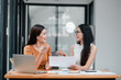 © Satori Studio - Two women smiling and talking at a desk in a modern office setting, with a laptop and paperwork.