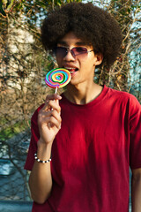 Dark-skinned Latino boy with braces and afro hair and sunglasses enjoying a colorful candy outdoors. Smiling, showing his braces and very happy.