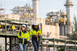 © kamonrat - Two female engineers in high-visibility jackets and hard hats conducting a field inspection at an industrial plant with refinery structures in the background, ensuring workplace safety and efficiency