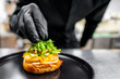 © pavel siamionov - A close-up of a hand in a black glove placing a gourmet burger with cheese and greens on a black plate, emphasizing freshness and quality. Perfect for culinary blogs, and restaurant promotions.