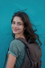 A woman stands in front of a bright blue wall, wearing a backpack and looking directly at the camera