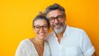 © Nadin Faust - portrait of a smiling middle-aged couple wearing eyeglasses, posing against a vibrant yellow background with copy space, exuding joy and fun while looking directly at the camera, isolated over a brigh