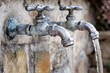 © Stanley - A close-up of an old metal faucet dripping water against a weathered wooden wall in a residential backyard during daylight