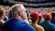 © ECLIVN - A senior man with gray hair and a white beard sits among a large crowd wearing a blue shirt and sunglasses. He's looking to his right, seemingly focused on an event in the distance.