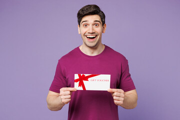 Young shocked surprised man wear violet t-shirt casual clothes hold gift certificate coupon voucher card for store isolated on plain pastel light purple background studio portrait. Lifestyle concept.