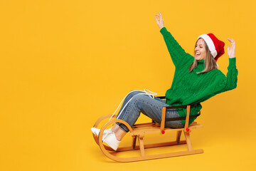 Wall Mural - Full body side view merry young woman wears green casual sweater Santa hat posing ride sled raise up hands isolated on plain yellow background. Happy New Year, celebration Christmas holiday concept.