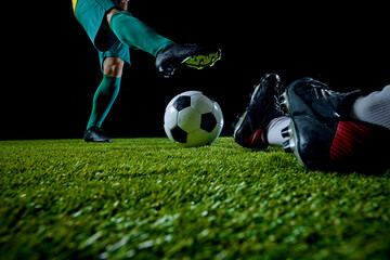 Close-up shot of soccer player in green uniform preparing to kick ball, while opponent on ground, on green football field against black studio background. Concept of sport event, match, championship