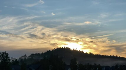 Wall Mural - Sun falls behind tree cover hillside with smoke and fog dancing below colorful clouds in sky at dusk.