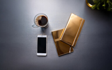 A minimalist flat lay of a smartphone, two gold notebooks, and a glass of coffee on a silver table.