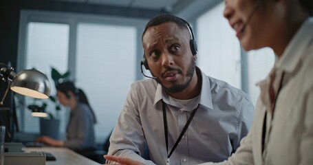 Call center office: Two multiethnic hotline operators in headsets discuss work, use computer having conversation with client on call. Technical support specialists working in online customer support.