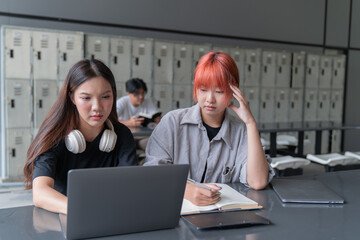 Two concentrated female students are working together on a project using a laptop and taking notes in a library with lockers in the background, showcasing collaborative learning