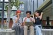 © Tj - Group of cheerful young students sitting together outdoors raising arms and celebrating success while looking at laptop, expressing excitement and achievement in academic or project accomplishment